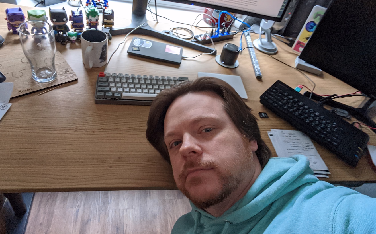 Remy with his head resting awkwardly on a desk, with a spectrum, a modern keyboard, some kind of phone and various figurines from lego Batman characters to super mini Jokers
